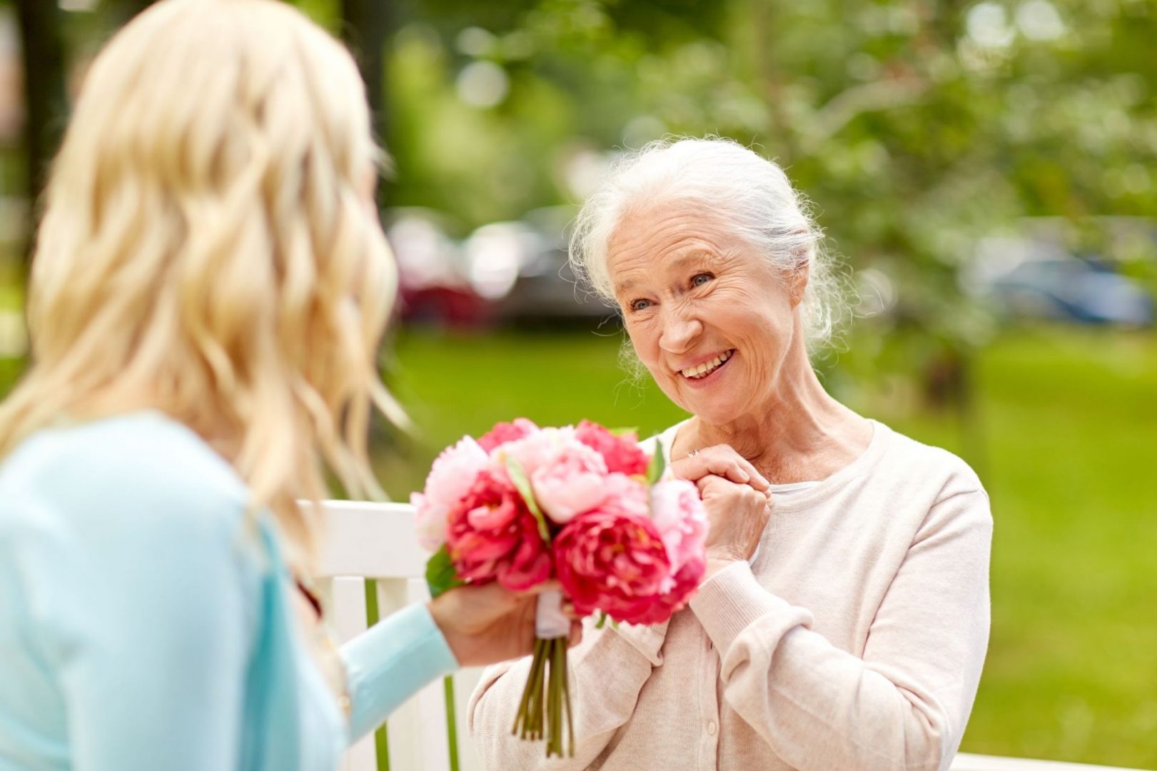 Senior woman smiling as she receives flower bouquet from young woman outdoors.