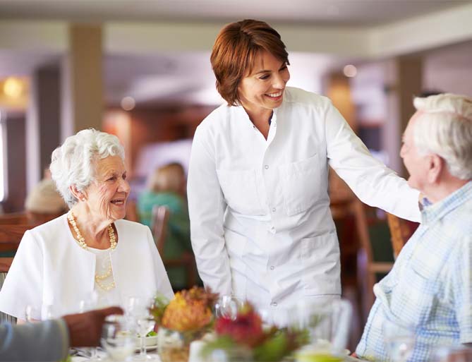 Caregiver smiles and talks with elderly residents in a dining area.
