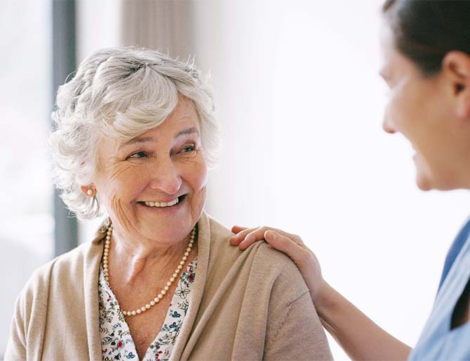 Elderly woman smiling with a caregivers hand on her shoulder in a senior living unit.