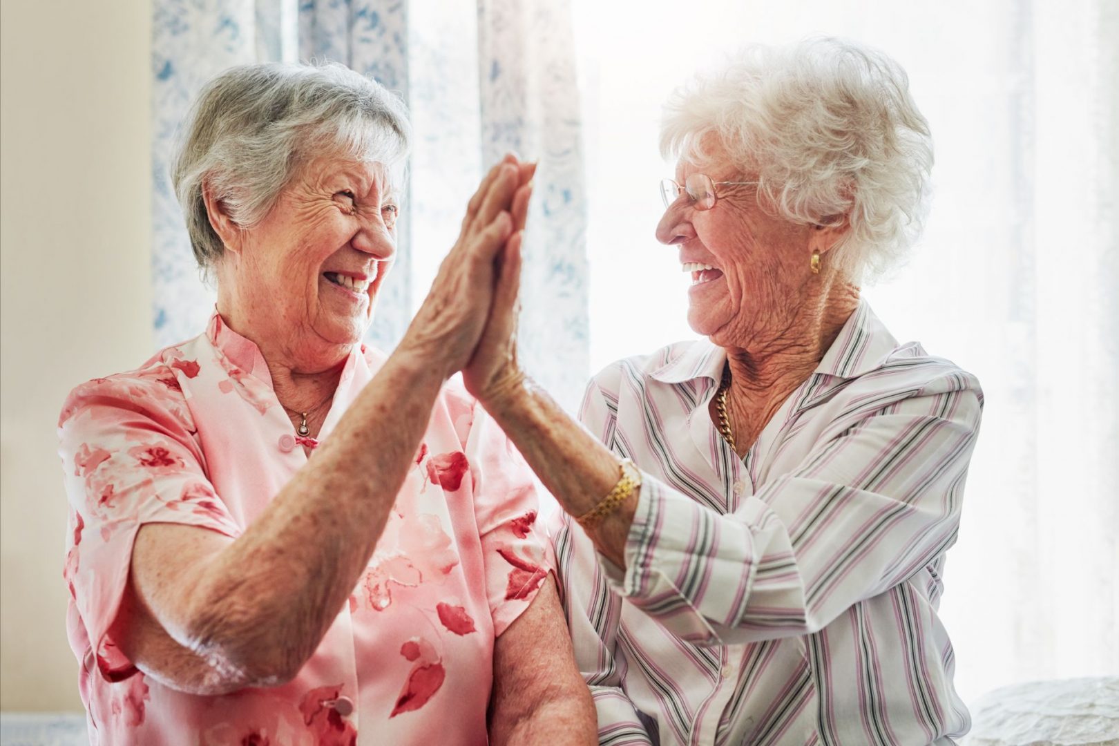 Two elderly women, smiling and giving a high-five in a bright, well-lit room with floral curtains.