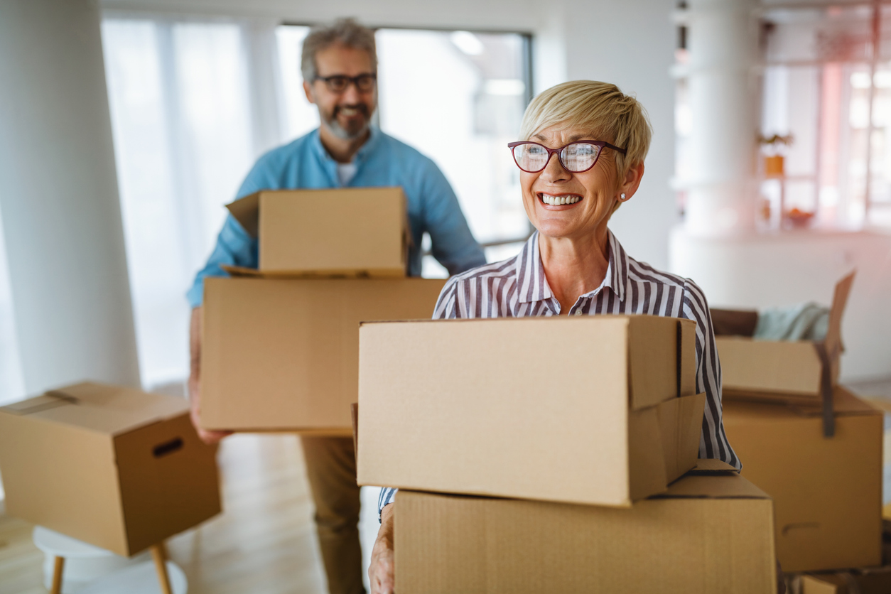 Smiling older couple carrying moving boxes into a bright room at their new residence.