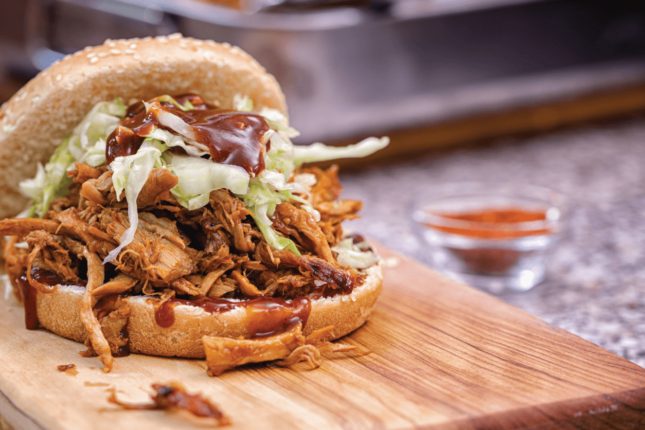 BBQ sandwich with pulled pork, coleslaw, and sauce on a wooden cutting board.