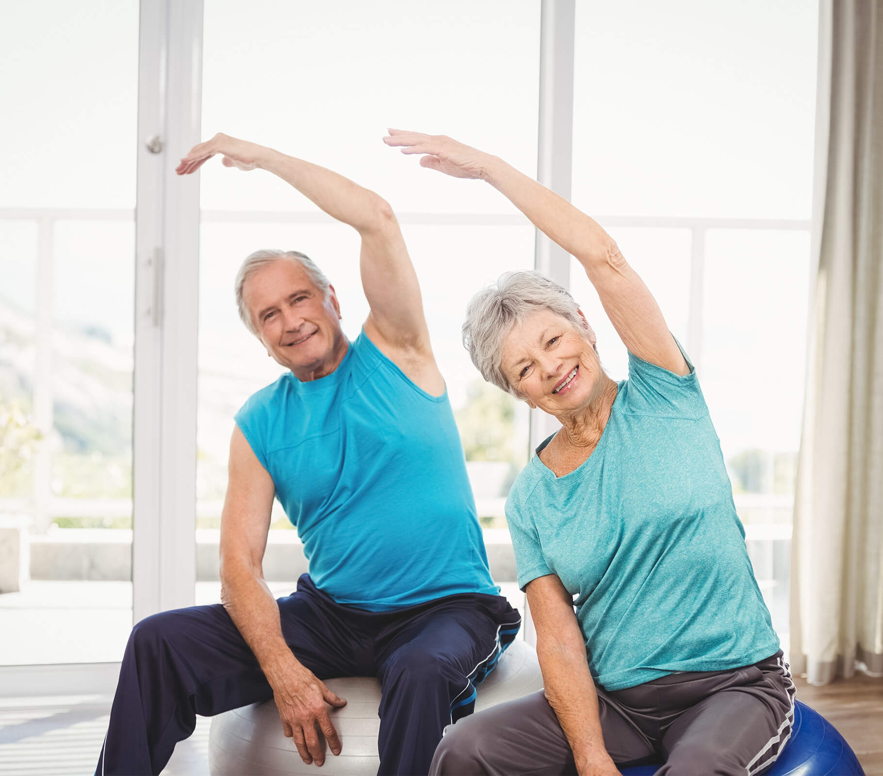 Elderly man and woman doing seated side stretches on exercise balls indoors.