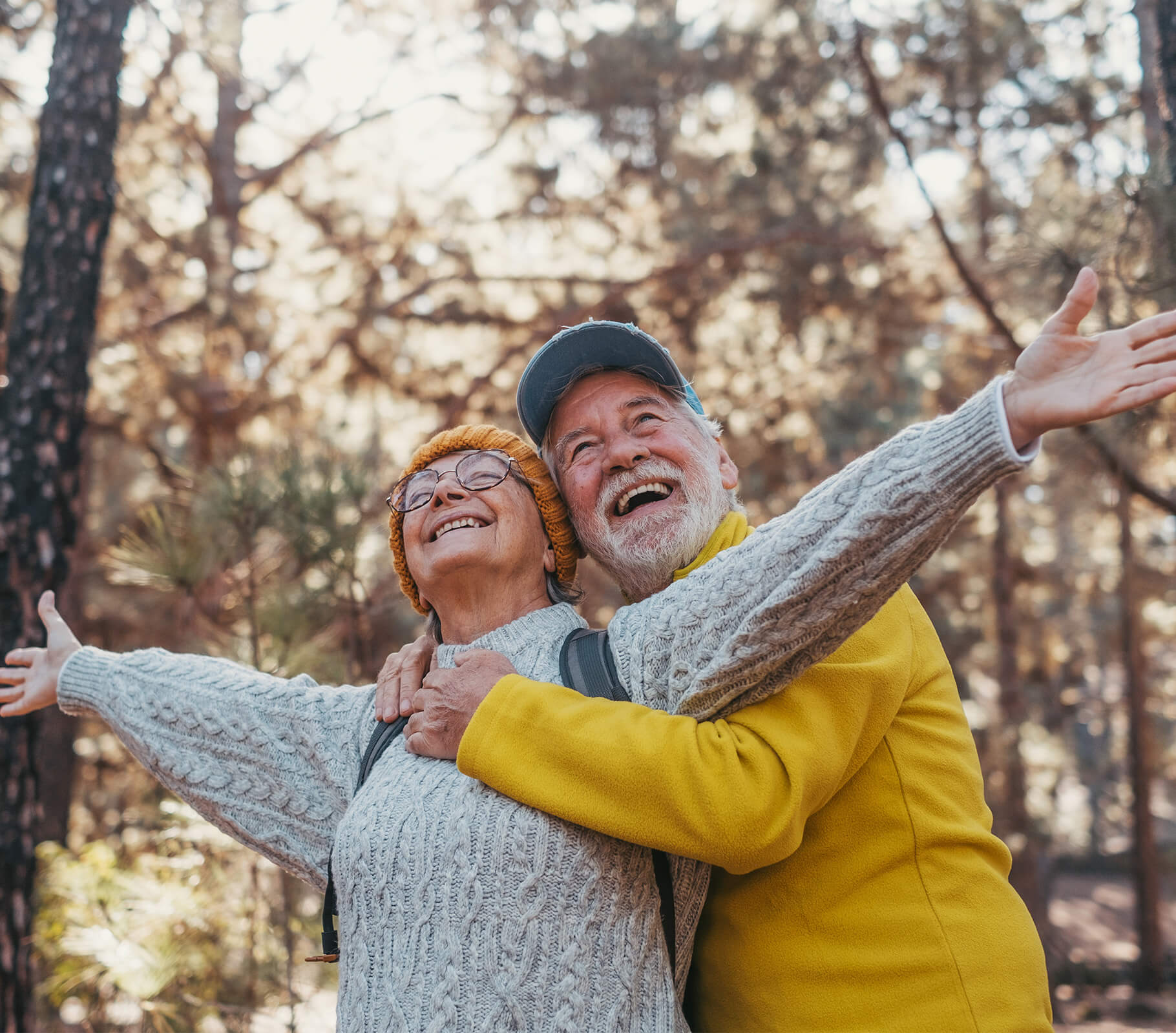 Elderly couple joyfully embracing in a sunlit forest, wearing cozy sweaters.