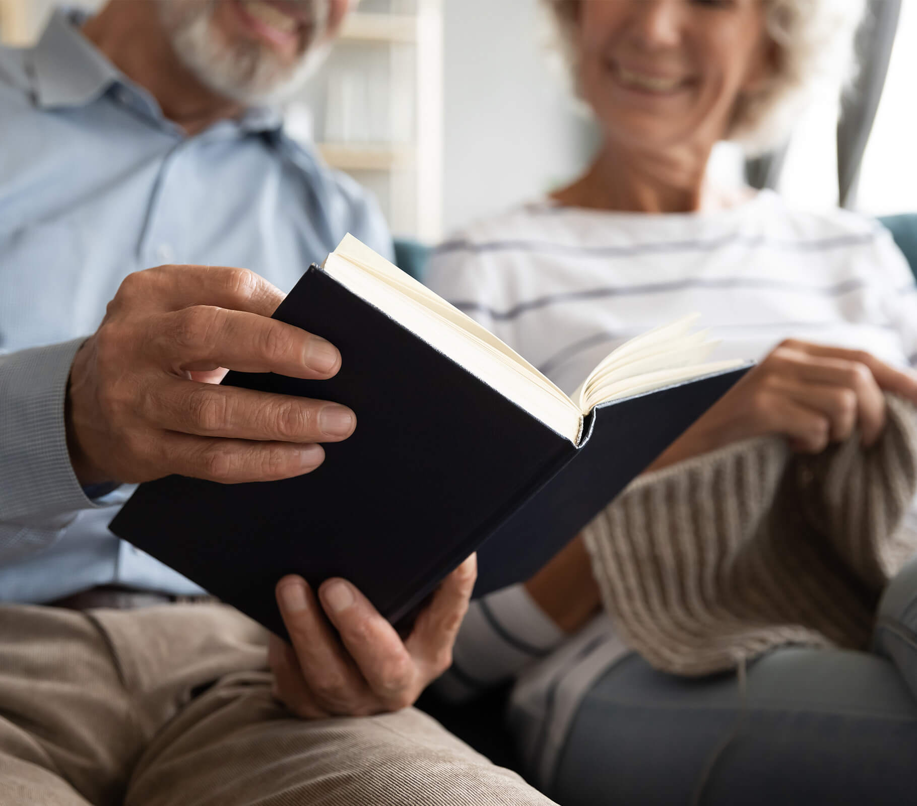 Elderly couple sitting on sofa reading a book together and smiling.
