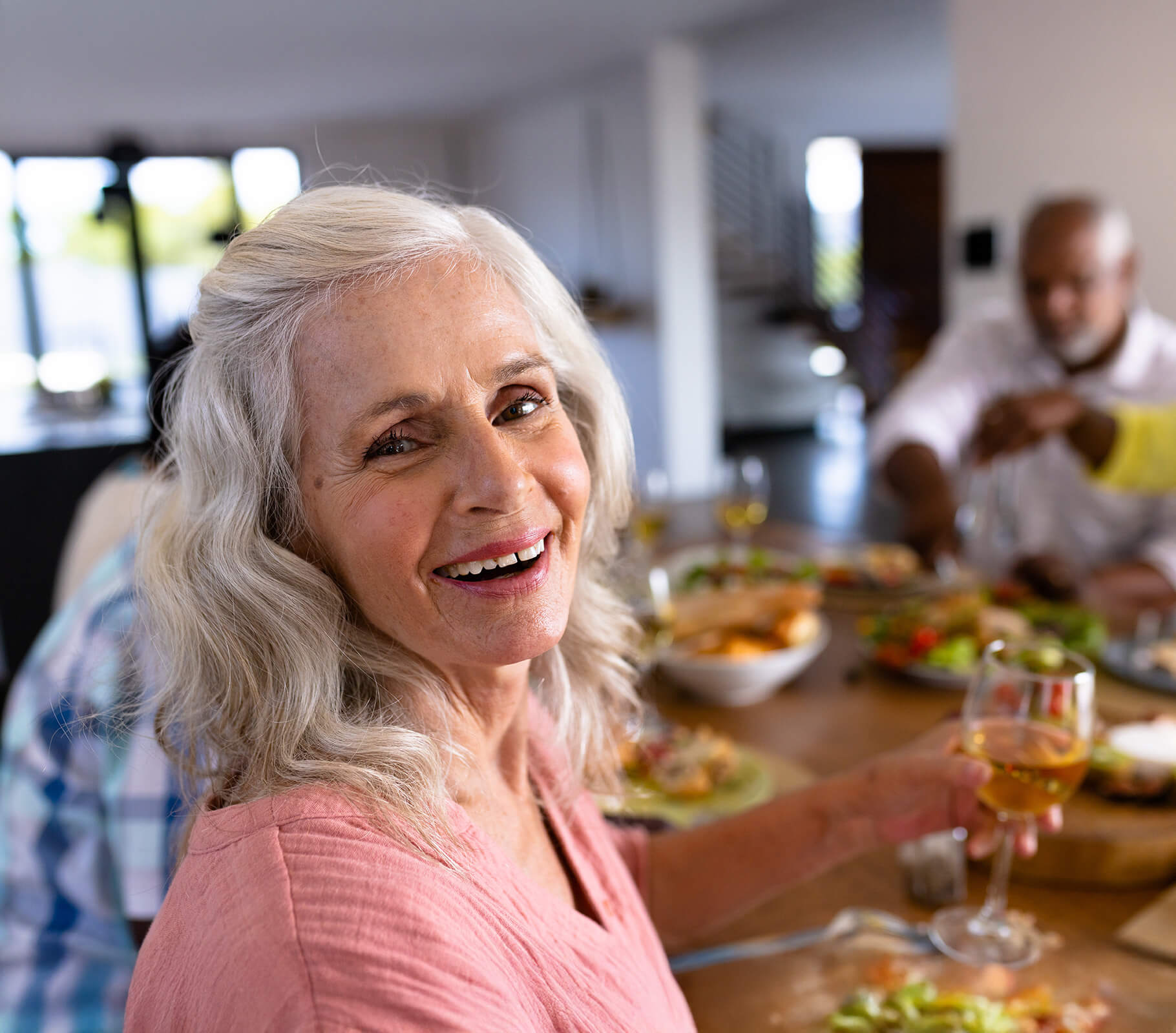 Elderly woman smiling at a communal dining table, holding a glass of wine.