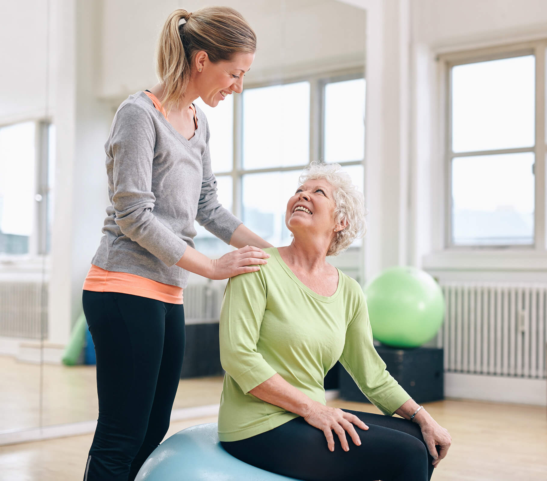 Woman assisting senior on exercise ball in a bright room with large windows.