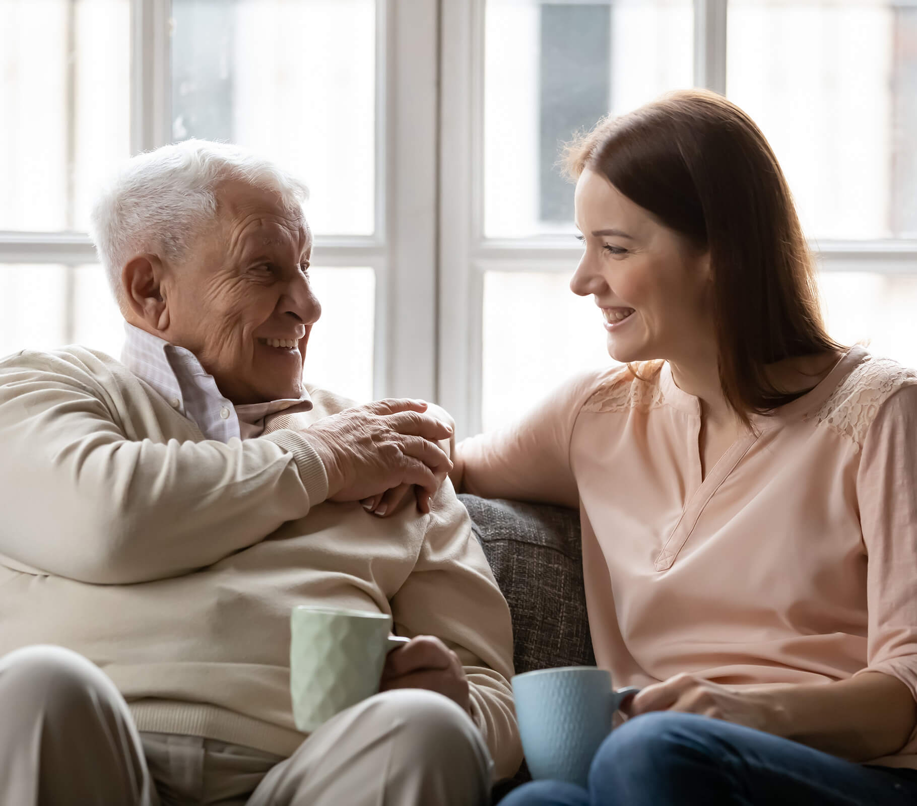 Elderly man and woman smiling, holding mugs, seated on a sofa in daylight.