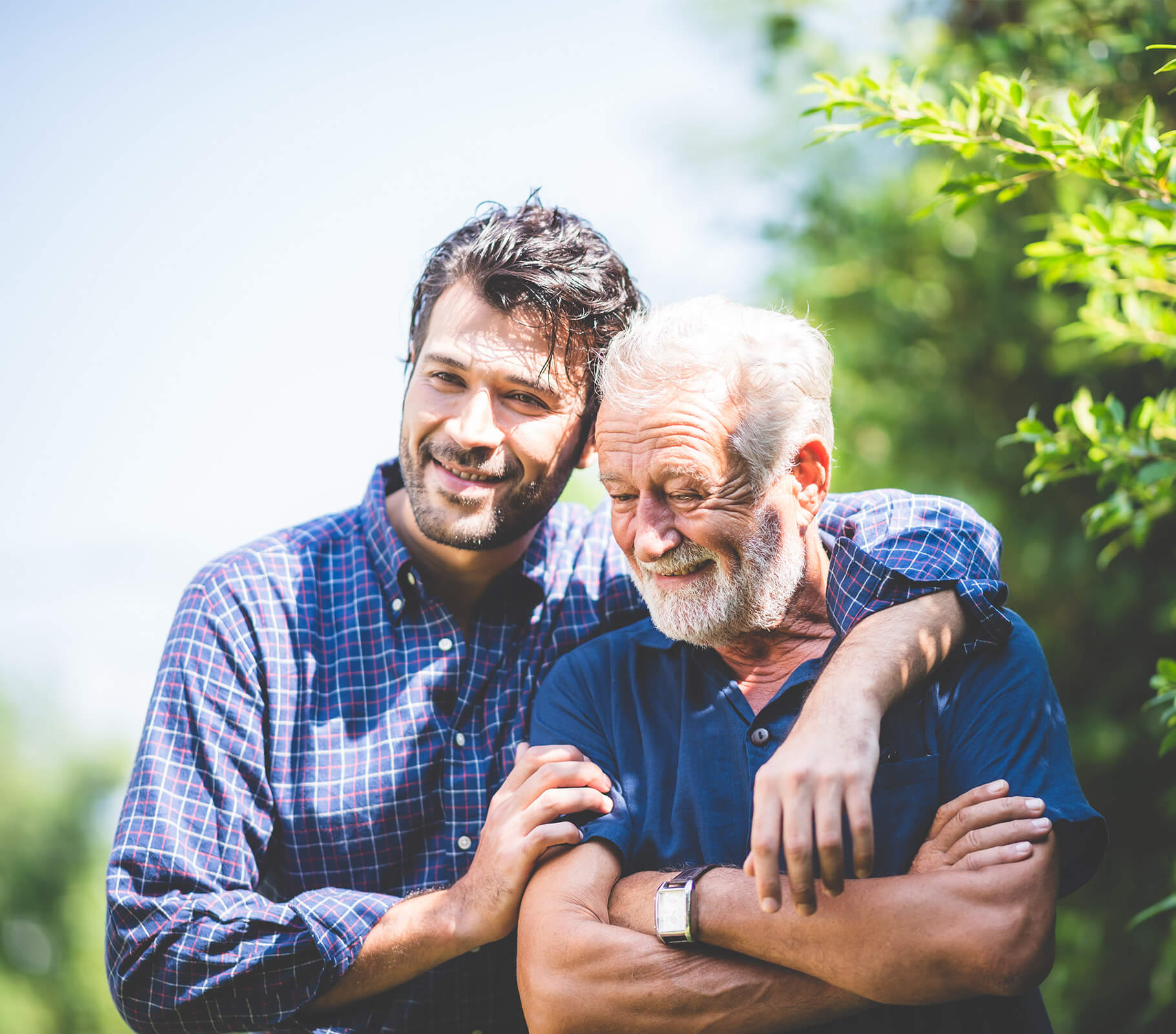 Younger man embraces older man, both smiling in a sunny garden setting.