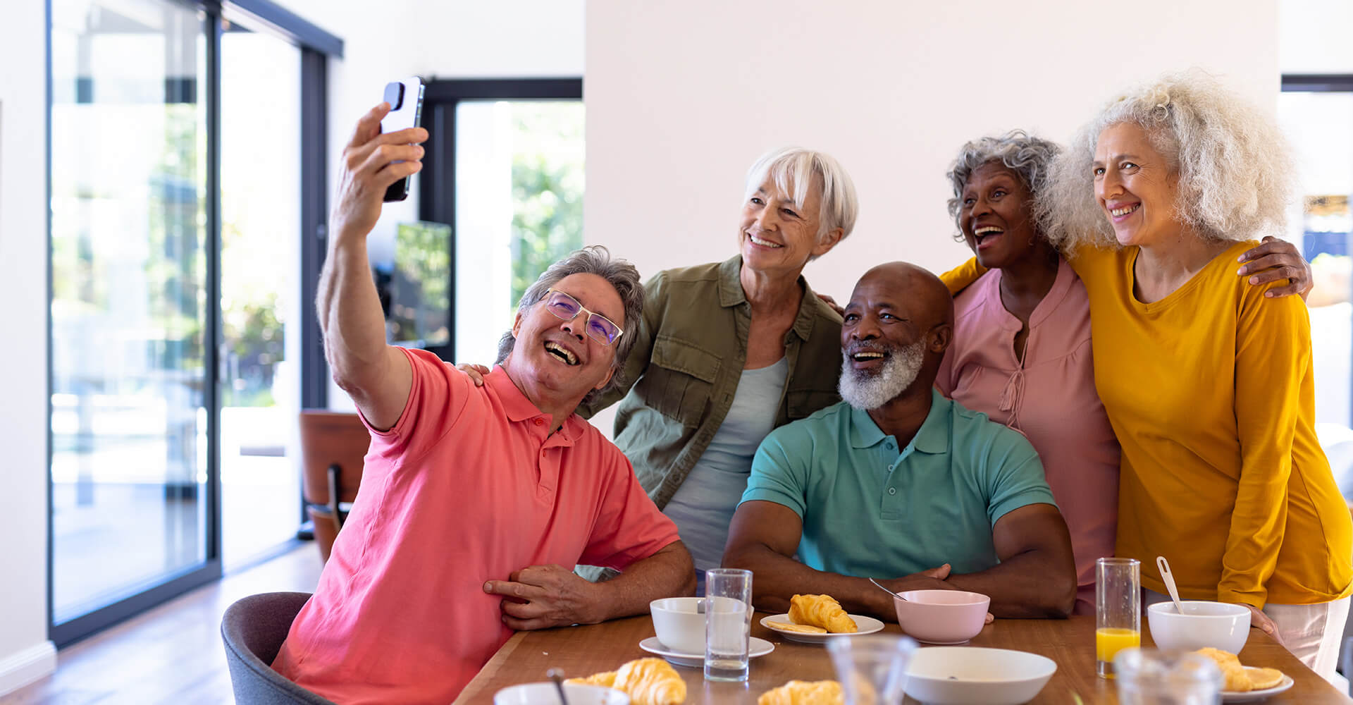 Group of seniors taking a selfie in a bright dining area with a wooden table.