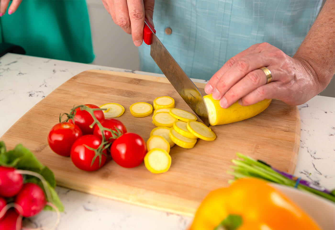 Person slicing yellow squash on a wooden board surrounded by fresh vegetables.