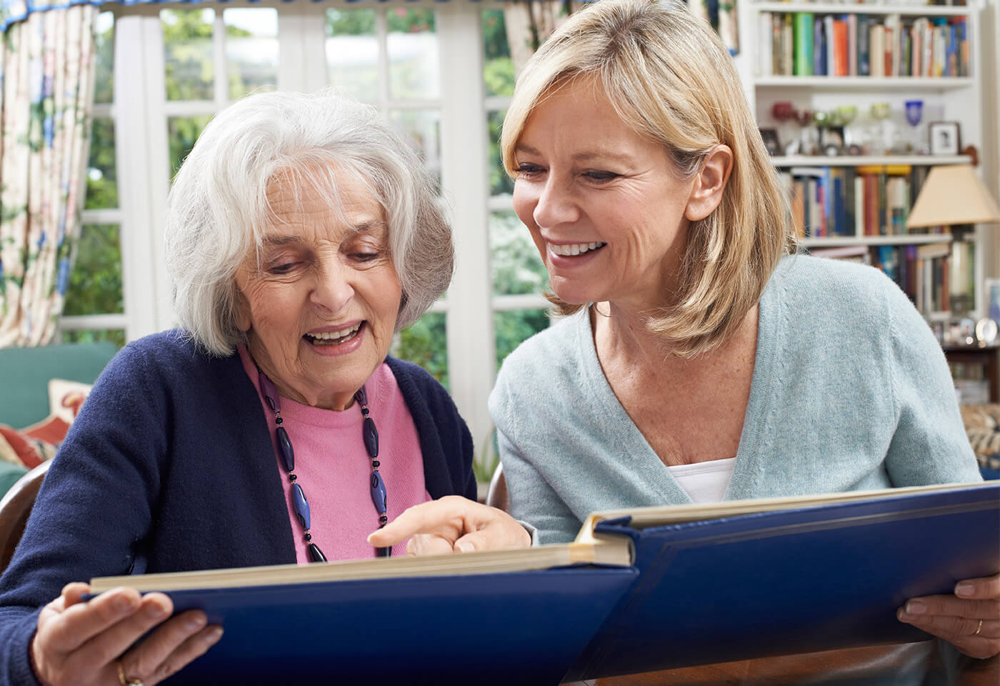Elderly woman and younger woman smiling while looking at a photo album in a cozy room.