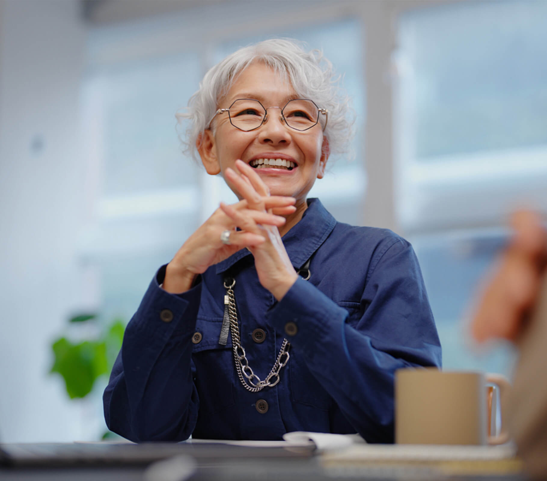 Elderly person with glasses smiling indoors, wearing a navy blue outfit.