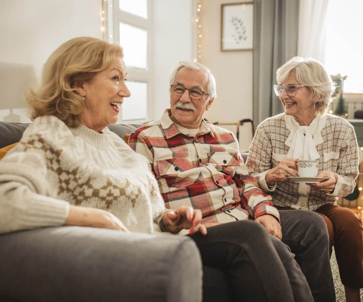Three seniors enjoying conversation in a cozy living room holding coffee cups.