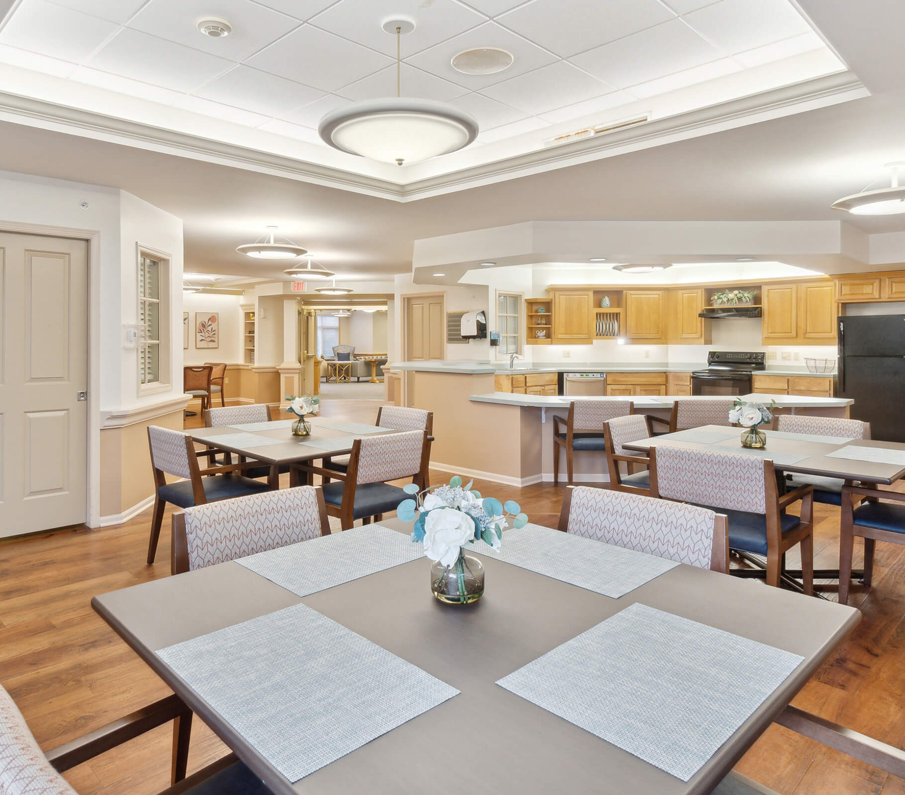 Well-lit dining area with tables, chairs, floral arrangements, and a modern kitchen.