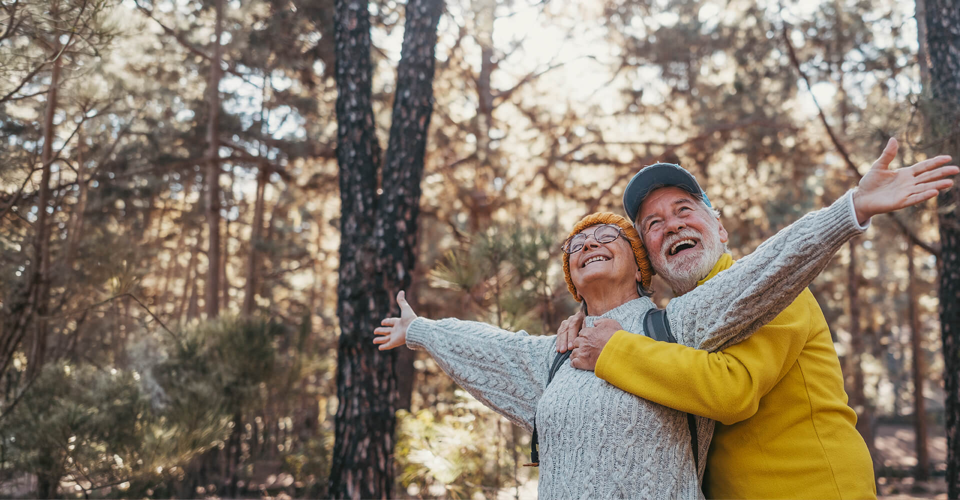 Elderly couple embracing joyfully in a forest setting, with arms outstretched.