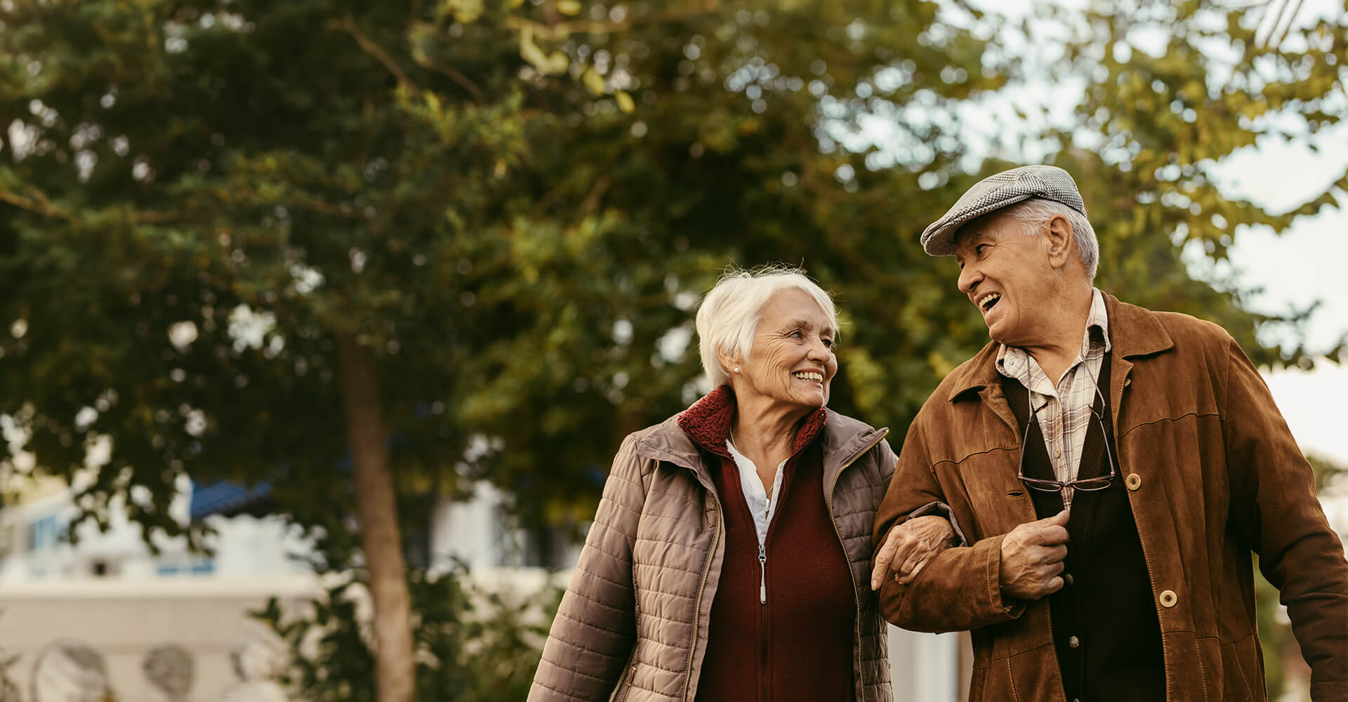 Elderly couple walking arm in arm through a tree-lined path in the community.