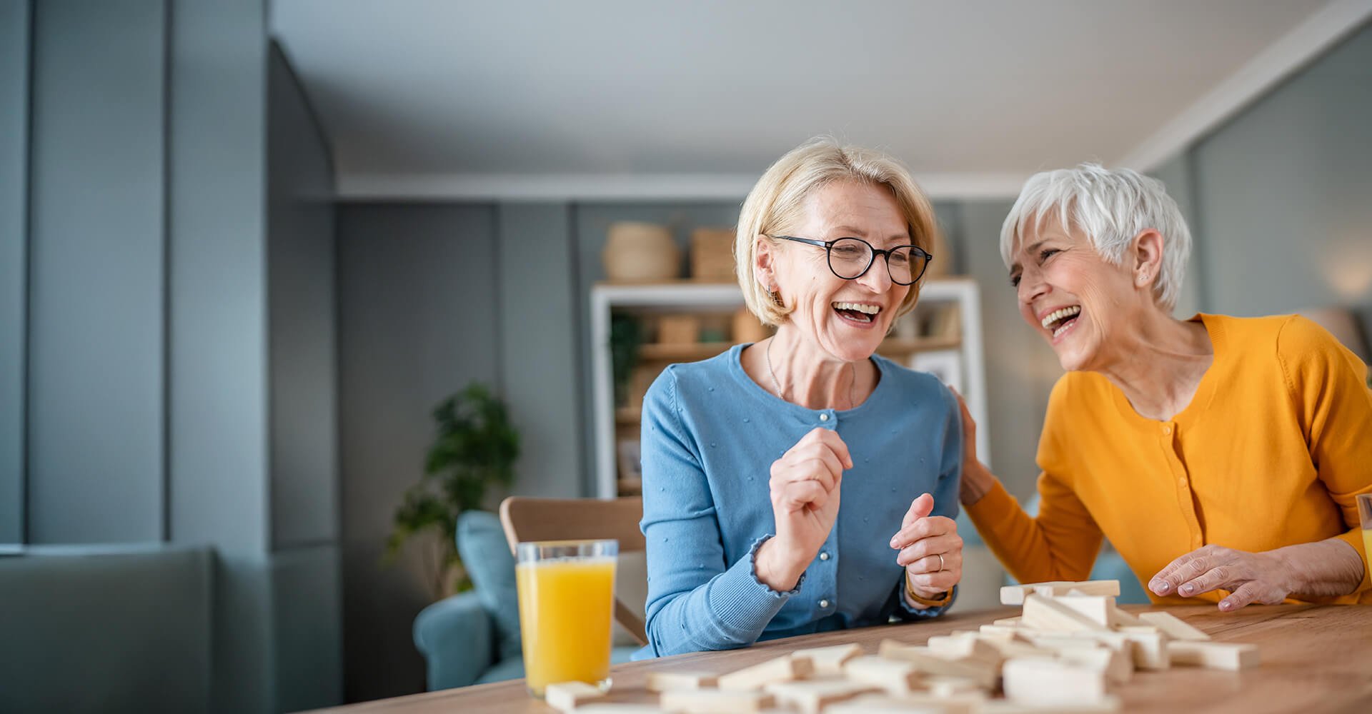 Two elderly women laughing and playing a block game at a wooden table.